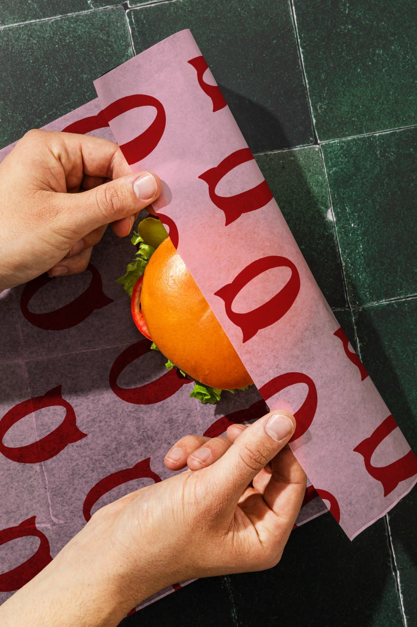 Руки заворачивают бургер в розовую бумагу с логотипом Q