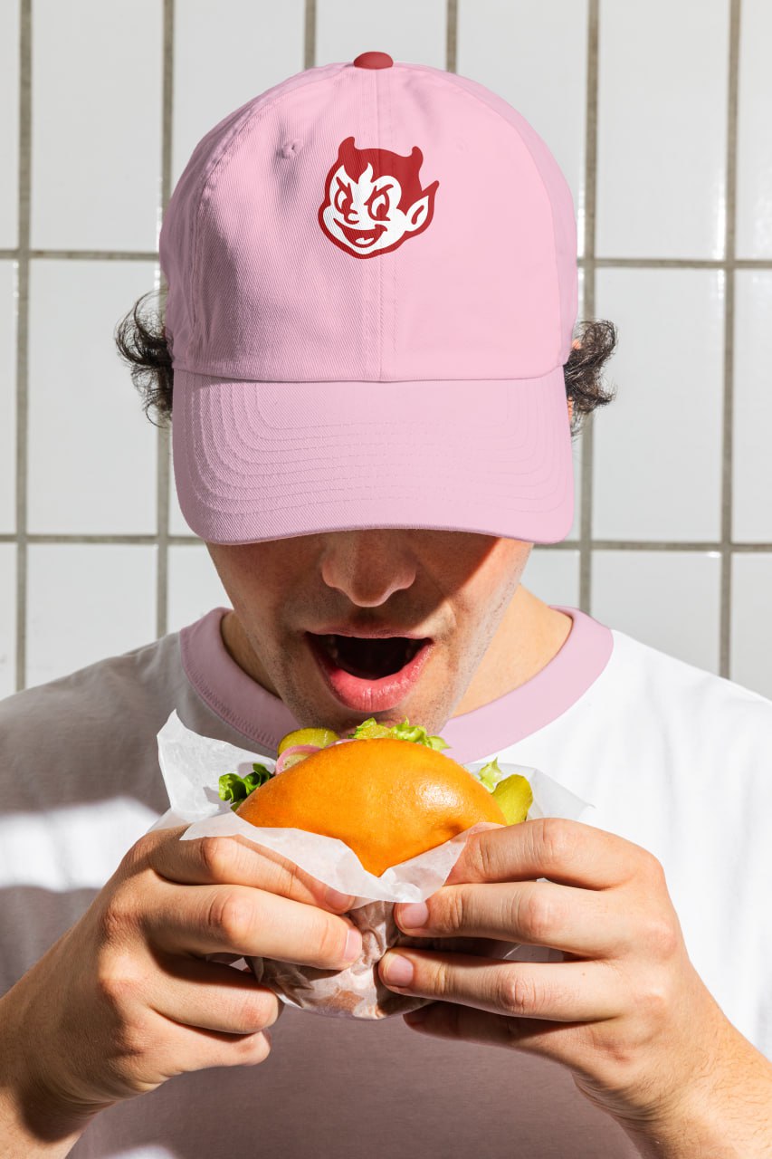 Молодой человек в розовой кепке с дьяволом собирается съесть гамбургер